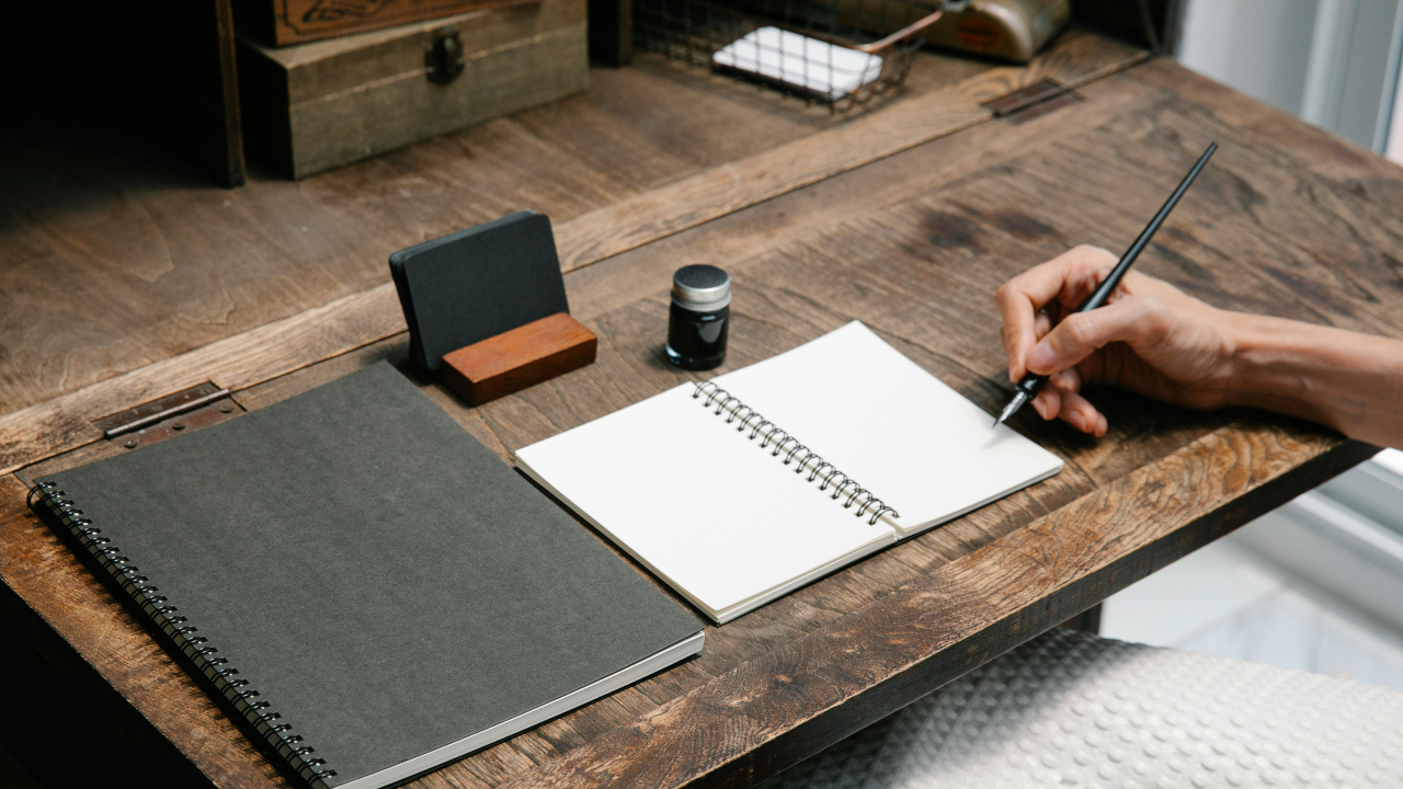 Девушка сидит за столом над раскрытой тетрадью, с пером в руке