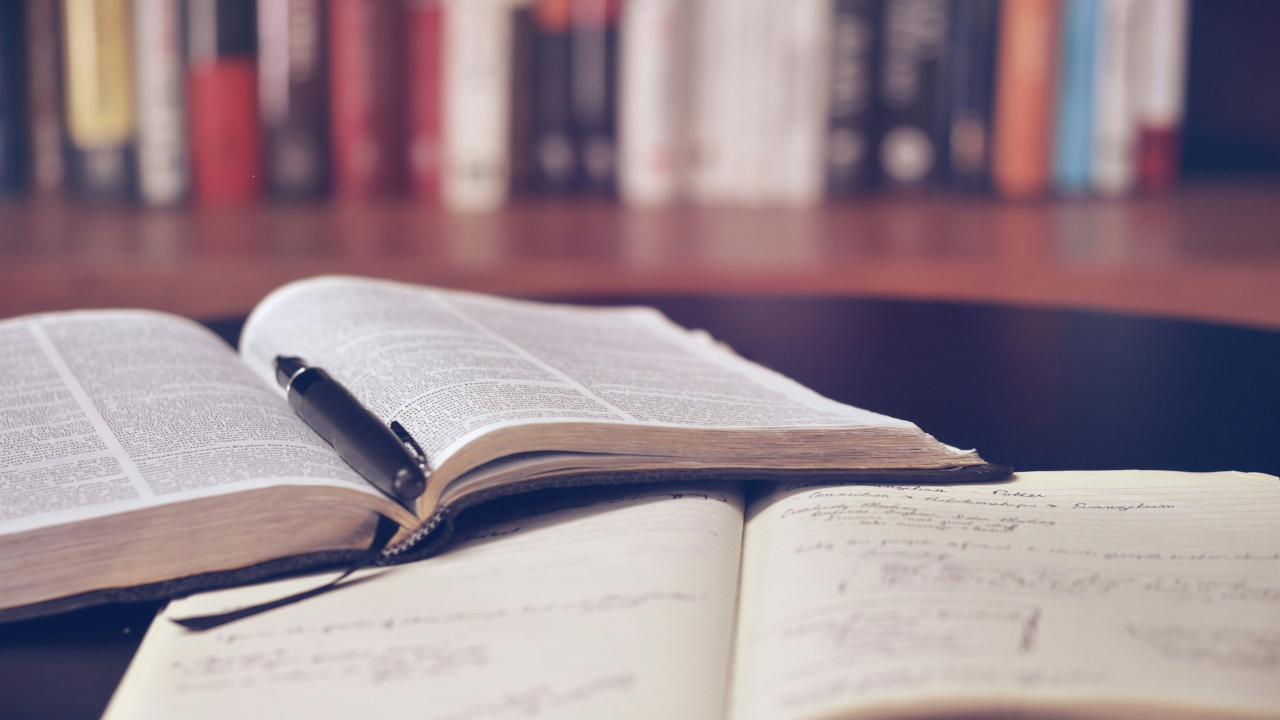 Раскрытая книга с ручкой и тетрадкой лежат на столе, на фоне стеллажа с книгами