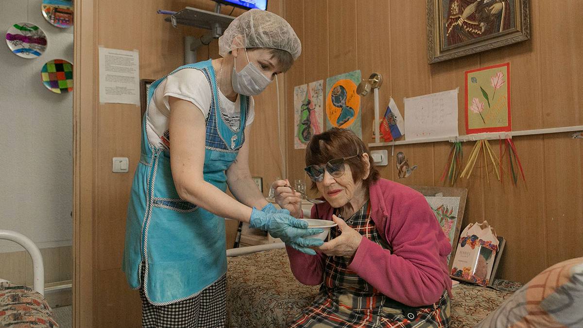 Женщина в фартуке помогает обедать пожилой пациентке